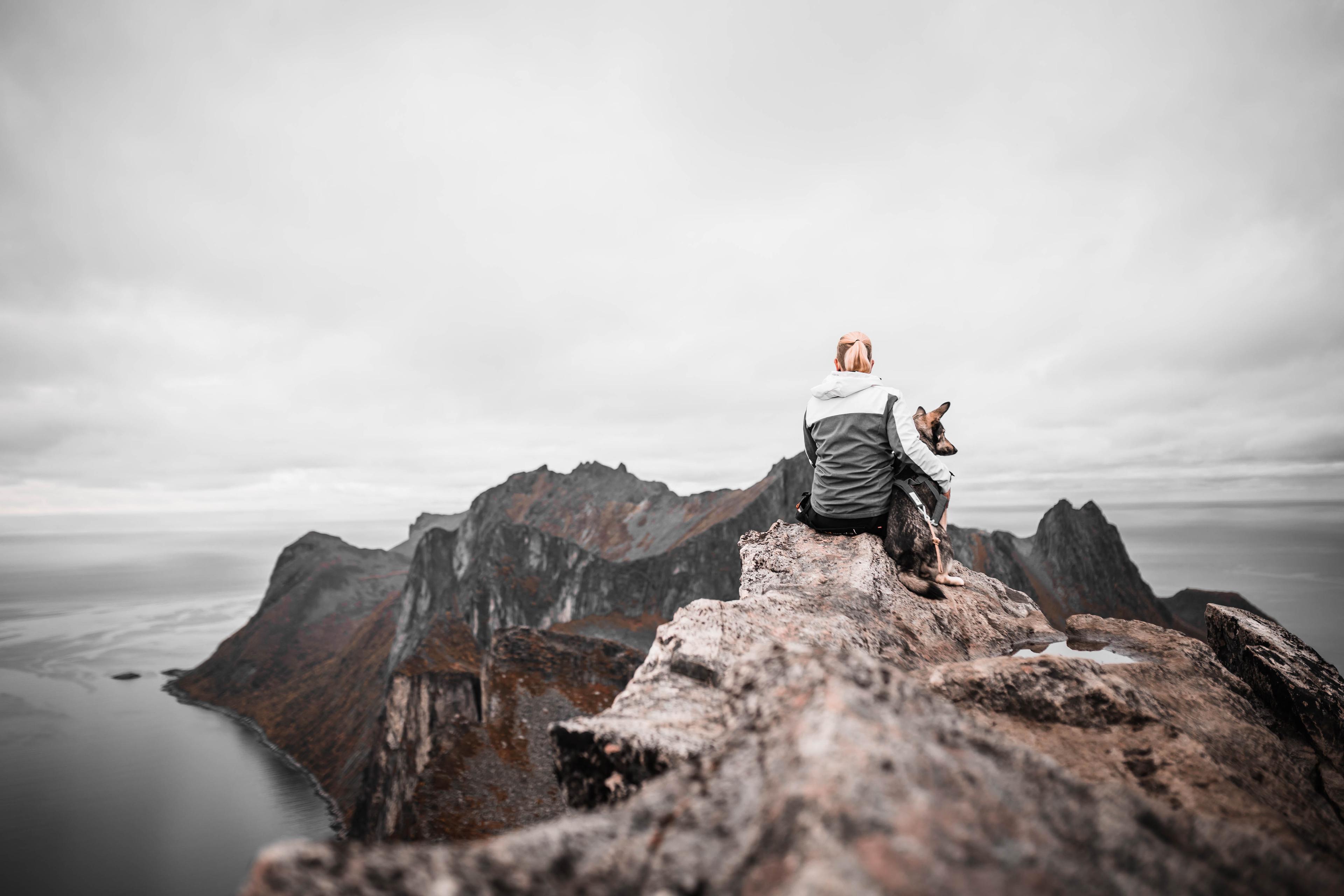 Anne and her dog sitting on a mountain ridge, facing the wild terrain together.